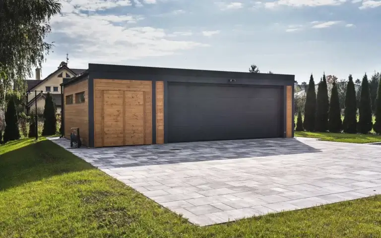 Na zdjęciu widzimy nowoczesny, wolnostojący garaż o eleganckim, minimalistycznym designie. Budynek łączy ciepłe drewno z ciemnymi, grafitowymi elementami, tworząc wyważoną kompozycję materiałową. Po lewej stronie znajduje się drewniane drzwi wejściowe z pionowymi deskami i prostym uchwytem, a po prawej — szeroka, gładka brama garażowa w ciemnym kolorze, nad którą zamontowano dwa symetryczne kinkiety. Garaż otacza przestronny podjazd z jasnoszarych płyt ułożonych w geometryczny wzór, a po bokach rozciąga się zadbany trawnik.