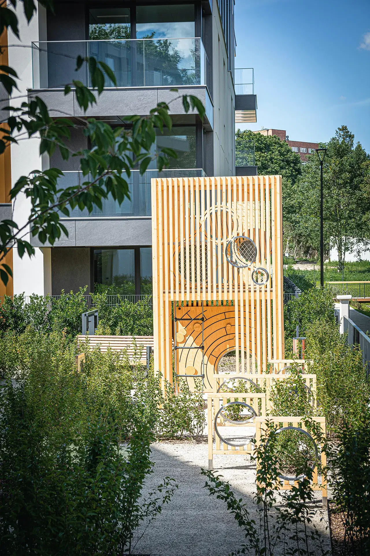 Na pierwszym planie znajduje się nowoczesna, drewniana konstrukcja placu zabaw z pionowymi panelami i geometrycznymi wycięciami, zaprojektowana z myślą o dzieciach. Wbudowane elementy wspinaczkowe i interaktywne panele zachęcają do ruchu i zabawy na świeżym powietrzu. Całość otoczona jest zadbaną zielenią oraz utwardzoną ścieżką.