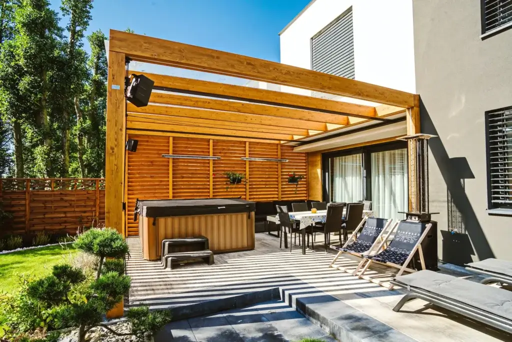 Nowoczesna pergola z jasnego drewna tworzy funkcjonalną strefę wypoczynkową przy domu, wyposażoną w stół z krzesłami, leżaki oraz jacuzzi. Przeszklone zadaszenie i drewniane lamele zapewniają ochronę przed słońcem.