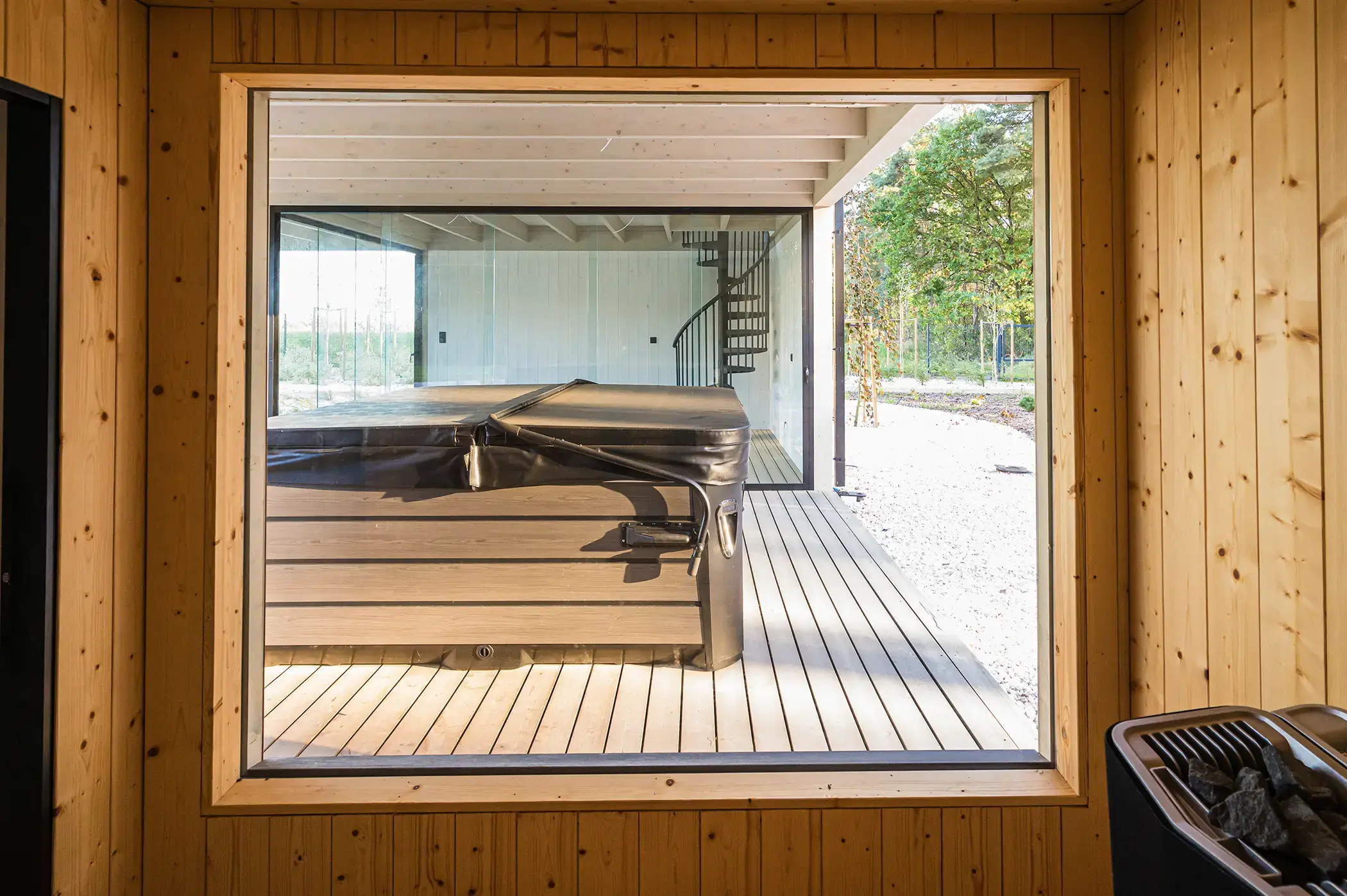 Na zdjęciu widać widok z wnętrza drewnianej sauny przez duże panoramiczne okno. Na zewnątrz, na drewnianym tarasie, znajduje się ogrodowe jacuzzi z pokrywą ustawione pod zadaszoną częścią budynku. W tle widać przeszkloną ścianę oraz metalowe schody spiralne prowadzące na wyższą kondygnację.