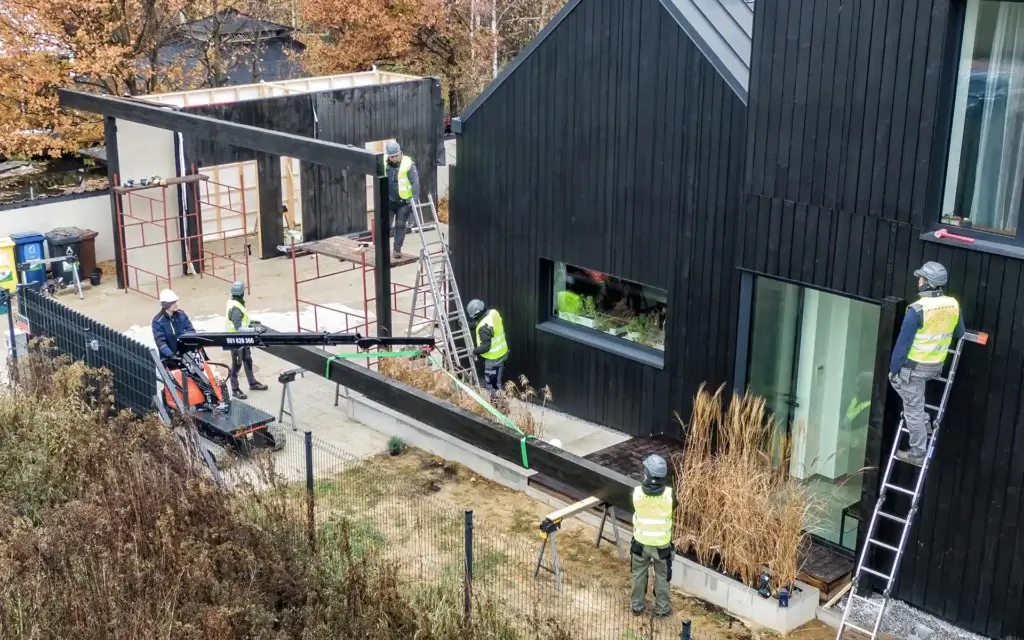 Na zdjęciu widać ekipę budowlaną pracującą przy montażu dużej, drewnianej belki konstrukcyjnej obok nowoczesnego domu z czarną, drewnianą elewacją. Belka jest podnoszona i ustawiana przy pomocy niewielkiego dźwigu, a kilku pracowników w kaskach i kamizelkach odblaskowych koordynuje jej ustawienie. Część osób stoi na rusztowaniach i drabinach, aby dokładnie dopasować element do konstrukcji. Wokół widać ogrodzenie posesji, podjazd oraz jesienną roślinność.