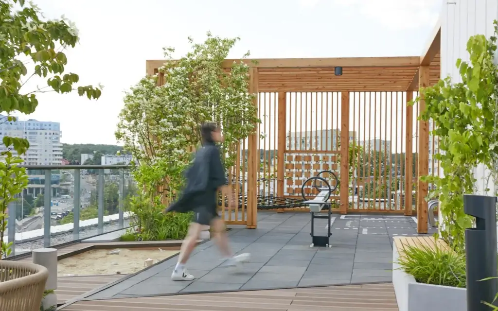 Na zdjęciu widać nowoczesny taras na dachu budynku, zaaranżowany jako przestrzeń rekreacyjna. Drewniana pergola i liczne rośliny nadają miejscu przytulny, naturalny charakter mimo miejskiego otoczenia. Przez kadr przechodzi osoba w ruchu, co dodaje scenie dynamiki. W tle widać wysokie budynki, które podkreślają, że jest to zielona oaza pośród miejskiej zabudowy.