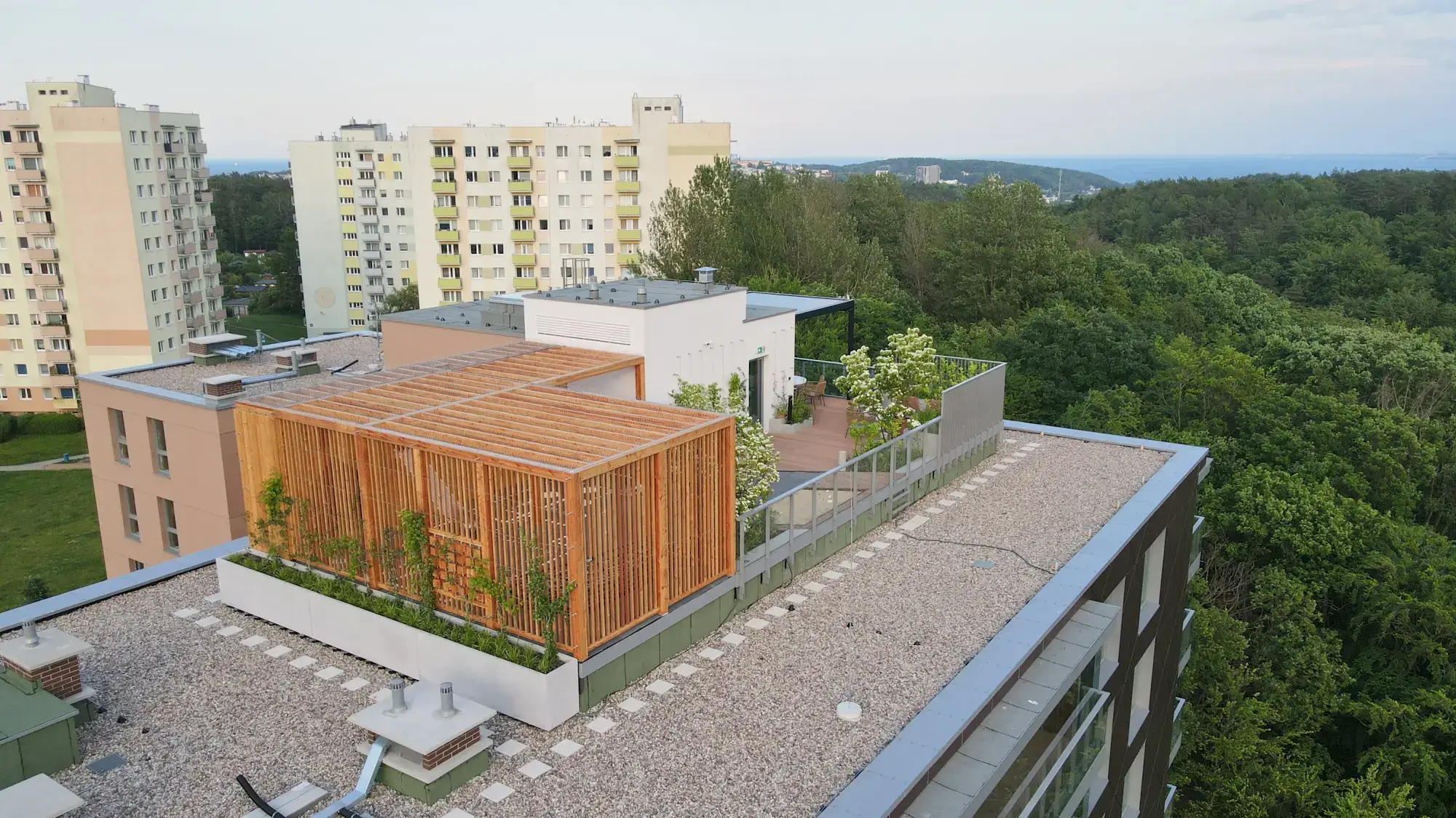 Na zdjęciu widać nowoczesny taras na dachu budynku, zaaranżowany jako elegancka przestrzeń wypoczynkowa. Drewniana pergola tworzy przytulne, częściowo zacienione miejsce z roślinami w donicach i miejscem do siedzenia. Podłoże pokryte jest żwirem, a przez całość prowadzą geometryczne płyty tworzące ścieżkę. W tle rozciąga się panorama miasta z wysokimi blokami, a za nimi widać las i daleki horyzont.