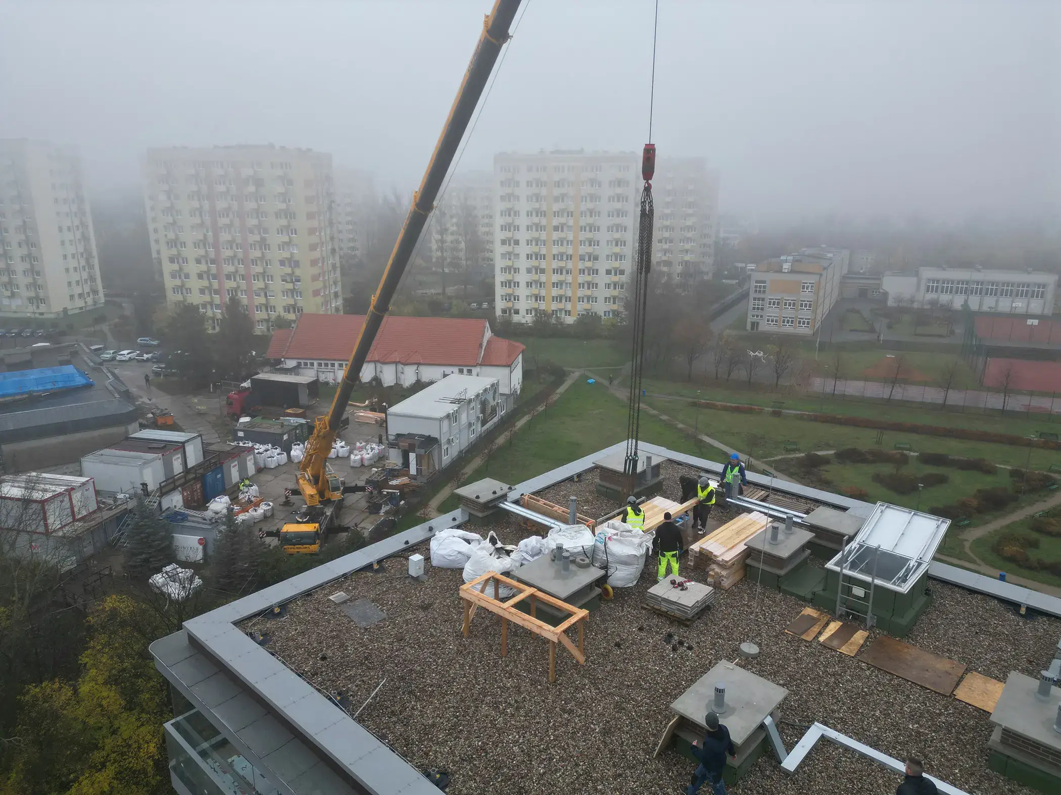 Na zdjęciu widać drewnianą konstrukcję w trakcie montażu, z pionowymi i poziomymi belkami tworzącymi szkielet przyszłej zabudowy. Prace odbywają się na dachu budynku, o czym świadczy widok na okoliczne bloki oraz wysokość, z której wykonano ujęcie. Na podłożu z płyt OSB leżą narzędzia i materiały, a w tle widać żółty dźwig podnoszący kolejne elementy. Kilku pracowników w odblaskowych kamizelkach zajmuje się organizacją materiałów, co podkreśla dynamiczny charakter budowy.
